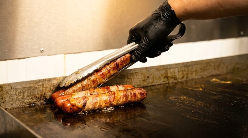 A gloved hand uses tongs to flip bacon-wrapped hot dogs cooking on a griddle.